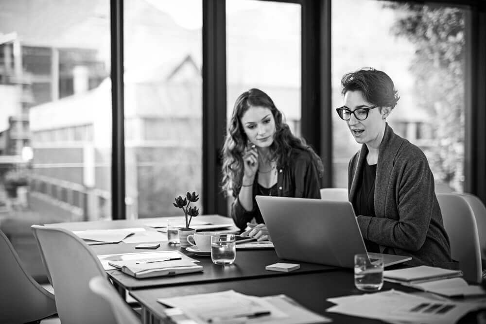 This is a photo of two business woman sitting at a desk looking at a laptop