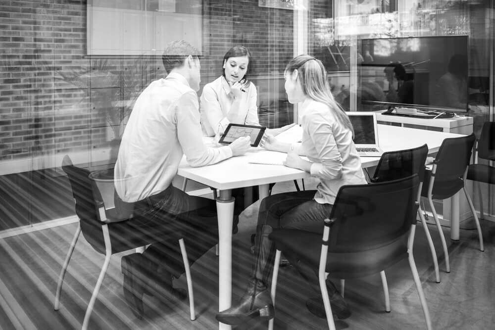 This is a photo of three people sitting around a desk looking at a monitor and talking